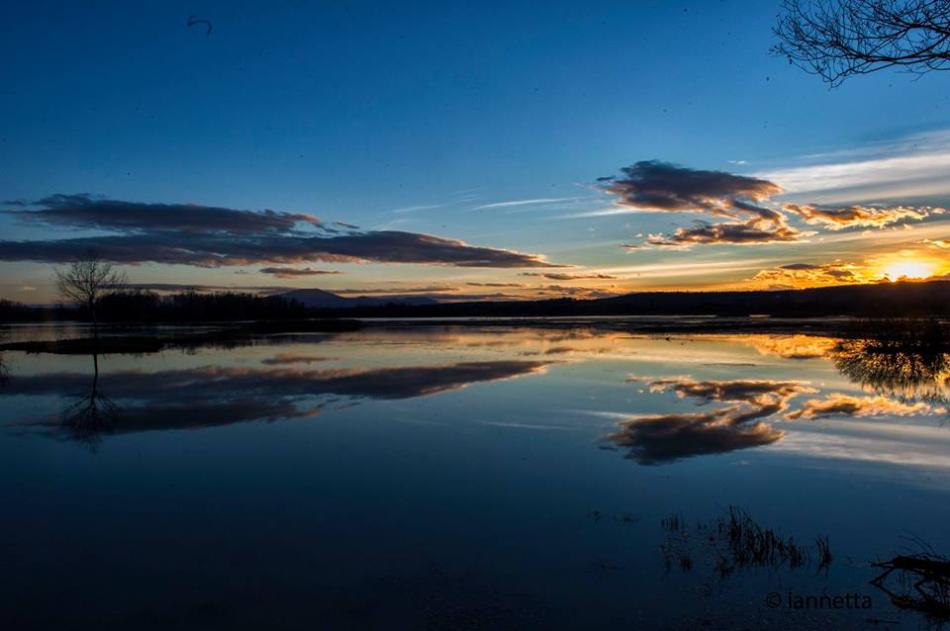Sunset over the lake with golden sky and clouds reflected in the calm waters, among silhouettes of trees and dark hills.